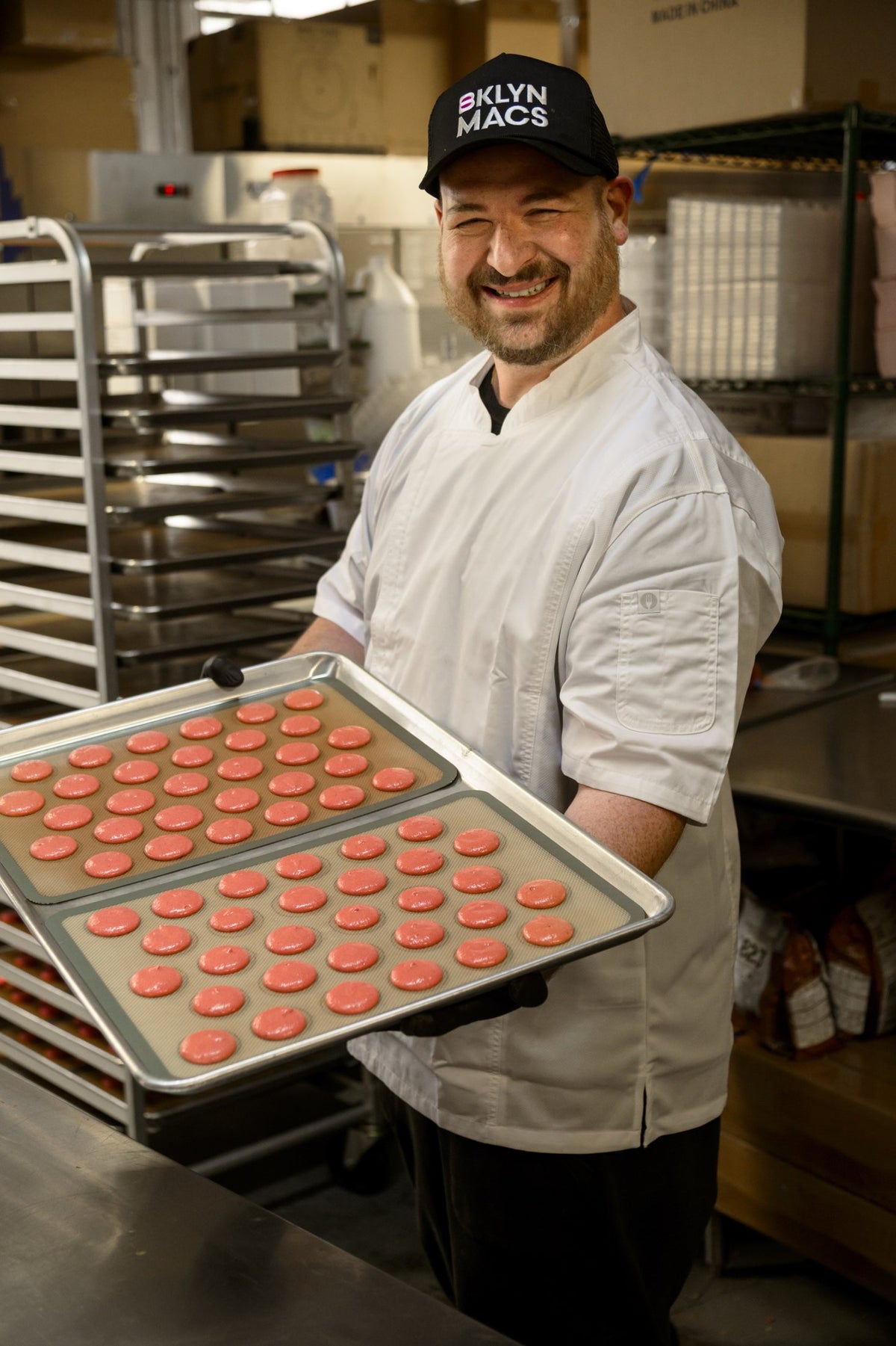 Gene smiling and holding two trays of freshly piped pink macaron shells
