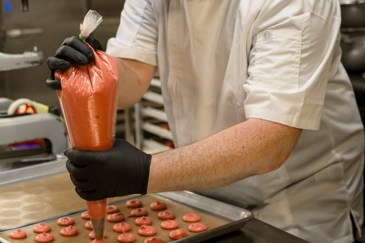 Gene piping rounds of pink macaron batter onto a lined baking tray