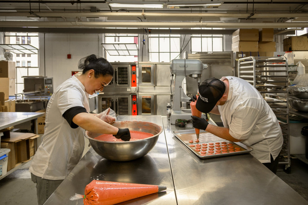 Rebecca folding macaron batter by hand while Gene pipes shells onto a tray in the BKLYN MACS kitchen
