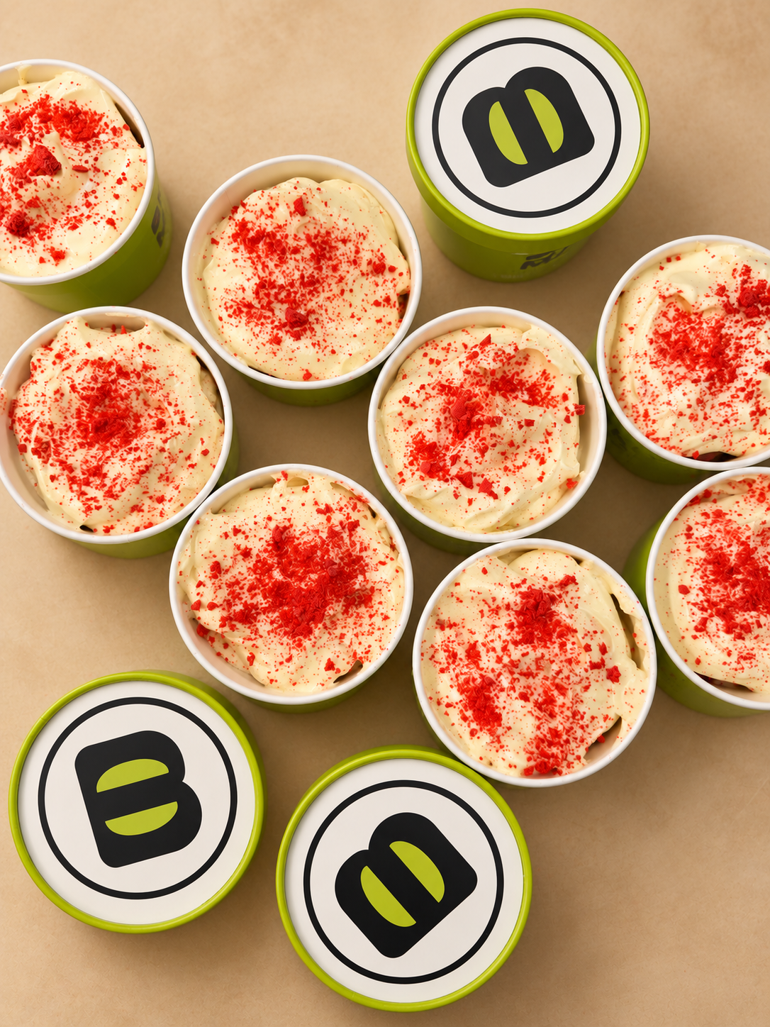 Cups of macaron banana pudding topped with cream and red crumbs, with lidded cups alongside