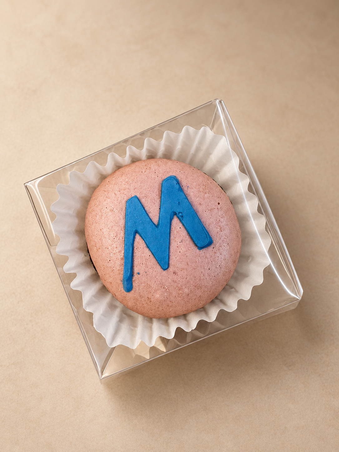 A single pink macaron with a raised blue Marshalls M logo standing up from the shell, in a clear favor box