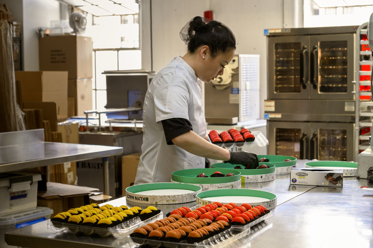 Rebecca filling BKLYN MACS gift tins with macarons, with trays of yellow, orange and red macarons in front